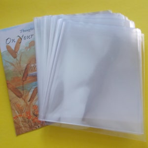 May include: A stack of clear, transparent plastic sleeves next to a book with the words "On Your" visible. The book has an illustration of reeds and a person. The background is a solid yellow.