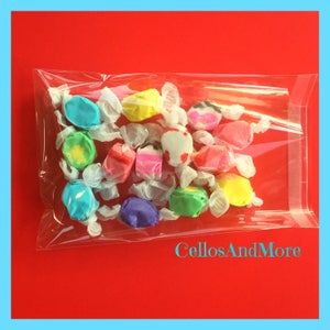 K&ouml;nnte beinhalten: Eine transparente Cellophant&uuml;te gef&uuml;llt mit bunten Salzwasser-Toffee-Bonbons. Das Toffee ist einzeln in wei&szlig;es Papier verpackt und in verschiedenen Farben erh&auml;ltlich, darunter Blau, Gelb, Rosa und Gr&uuml;n. Der Hintergrund ist rot mit dem Text "CellosAndMore".