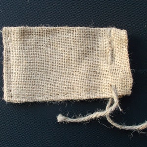 Op de afbeelding: Een kleine, rechthoekige jute zak in een natuurlijke beige kleur. De zak is gemaakt van geweven stof en heeft een eenvoudige, rustieke uitstraling. Een gedraaide touwstreng is aan de bovenkant bevestigd en dient als sluiting.