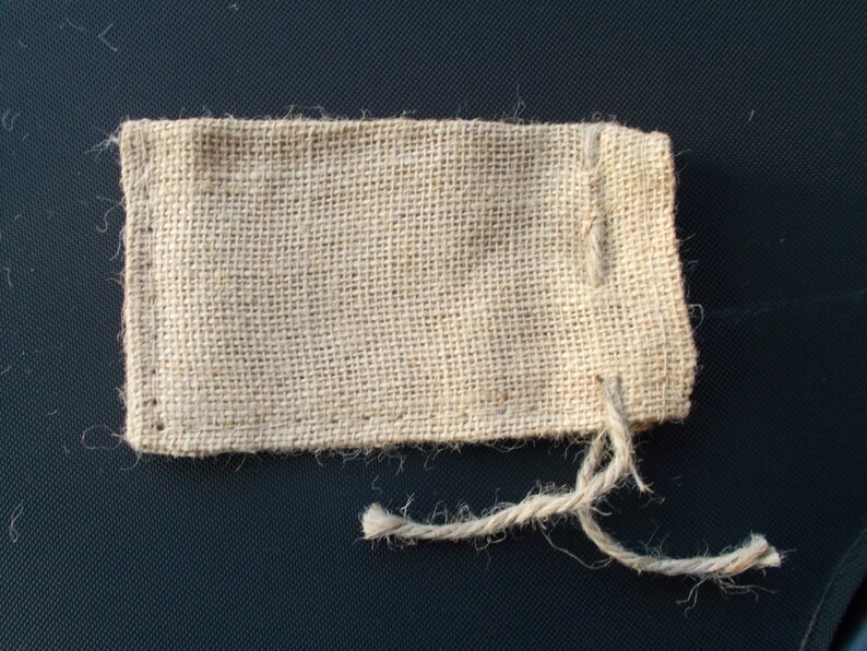 Op de afbeelding: Een kleine, rechthoekige jute zak in een natuurlijke beige kleur. De zak is gemaakt van geweven stof en heeft een eenvoudige, rustieke uitstraling. Een gedraaide touwstreng is aan de bovenkant bevestigd en dient als sluiting.