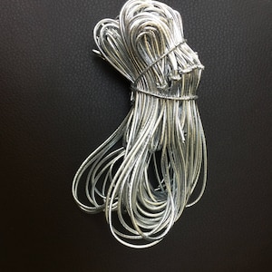 May include: A bundle of silver-colored cord, tightly bound in the middle, against a dark background. The cord appears to be made of a flexible material, with a slightly reflective surface. The bundle is neatly organized, with loops and curves.