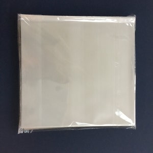 May include: A stack of clear, square, plastic sleeves, sealed in a transparent plastic wrap. The sleeves are a light, opaque color and are likely designed for storing and protecting items such as photographs or documents. The background is a dark blue surface.