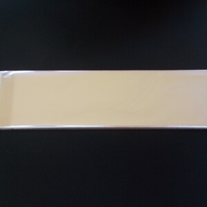 May include: A stack of rectangular, light tan, cardstock sheets, wrapped in clear cellophane. The stack is positioned horizontally against a black background. The cellophane is sealed on one end.