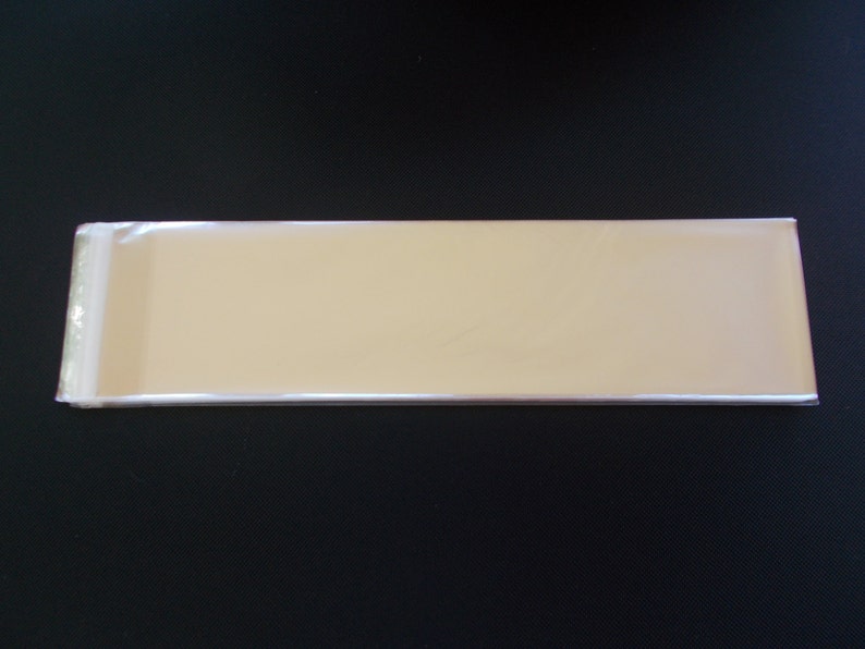 May include: A stack of rectangular, light tan, cardstock sheets, wrapped in clear cellophane. The stack is positioned horizontally against a black background. The cellophane is sealed on one end.