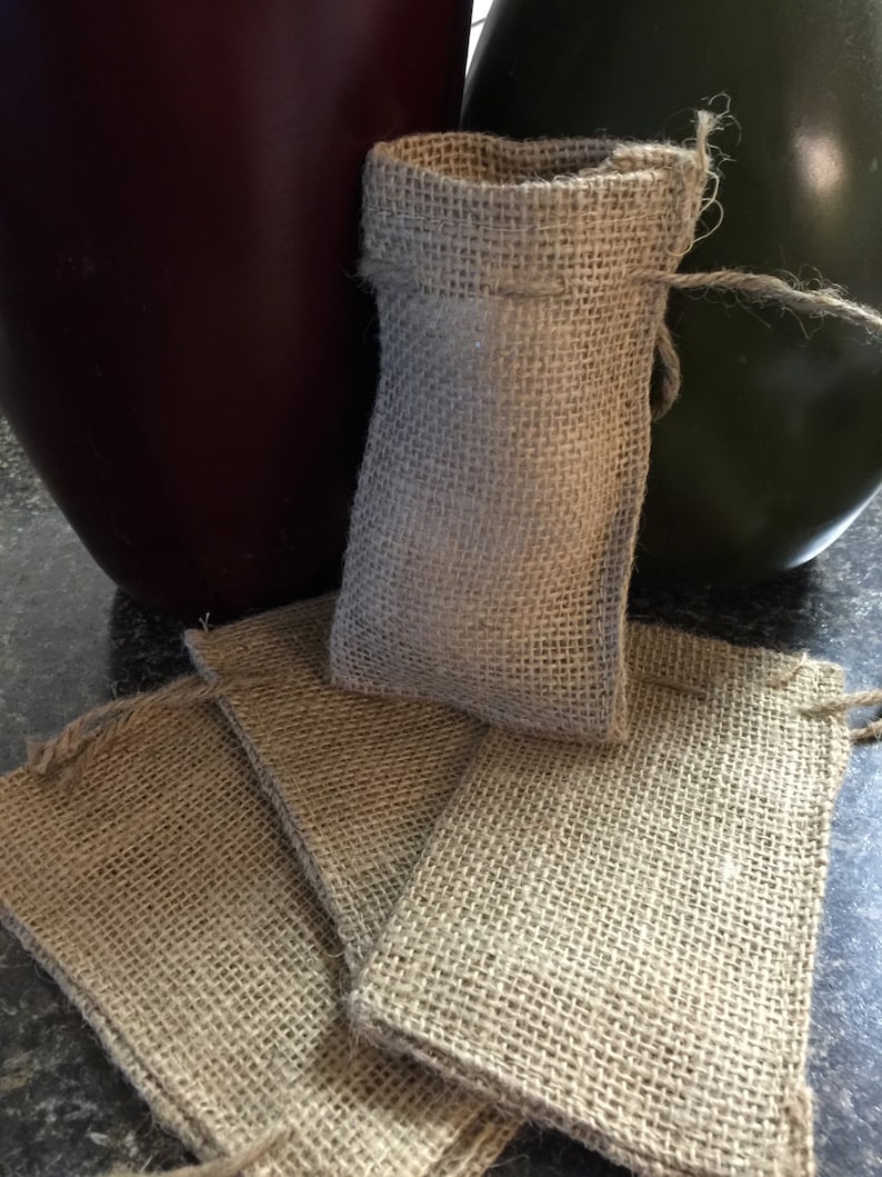 K&ouml;nnte beinhalten: Eine Sammlung kleiner Jutebeutel, einige flach liegend und andere aufrecht, mit Kordelzug. Die Beutel bestehen aus einem groben, gewebten Stoff. Ein dunkelrotes und ein gr&uuml;nes Dekorationsobjekt befinden sich im Hintergrund.