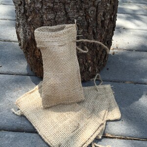Op de afbeelding: Een kleine, open jute zak met een trekkoordsluiting staat rechtop, met twee platliggende jute vierkanten. De items hebben een natuurlijke bruine kleur en zijn geplaatst op een grijze ondergrond, met een boomstam op de achtergrond.