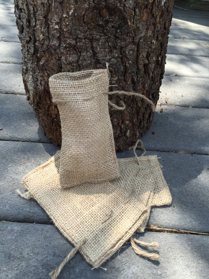 Op de afbeelding: Een kleine, open jute zak met een trekkoordsluiting staat rechtop, met twee platliggende jute vierkanten. De items hebben een natuurlijke bruine kleur en zijn geplaatst op een grijze ondergrond, met een boomstam op de achtergrond.