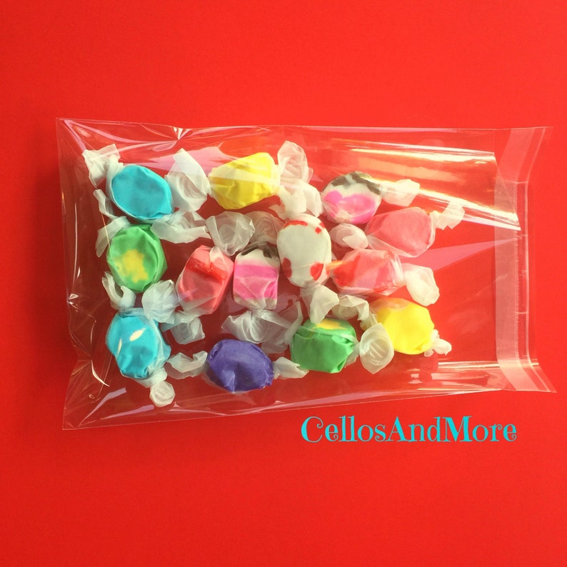 May include: A clear cellophane bag filled with colorful saltwater taffy candies. The taffy is wrapped in white paper and comes in various colors, including blue, yellow, pink, and green. The background is a solid red.