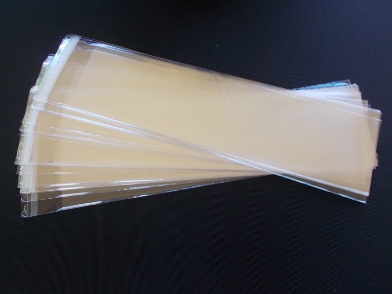 May include: A stack of clear, rectangular cellophane bags. The bags are transparent, allowing a glimpse of the tan-colored interior. The edges are sealed, and the bags are arranged in a fan-like pattern, suggesting they are ready for use.