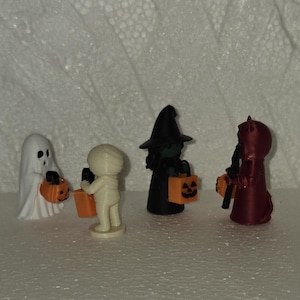 May include: Four small Halloween figurines standing in a row. From left to right, a white ghost, a beige mummy, a black witch with a pointed hat, and a dark red devil figure. Each character holds a small orange plastic pumpkin treat bucket.