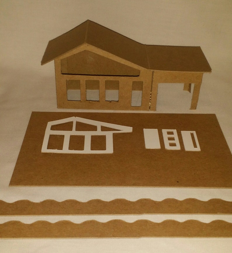 May include: A cardboard model house kit with a brown roof, windows, and a garage door. The kit also includes a separate piece for a sunroom and a wavy border.