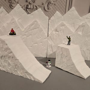 May include: White foam mountain backdrop with two figures, one sledding down a slope and the other standing on a peak. The backdrop is designed for miniature figures and model train sets.