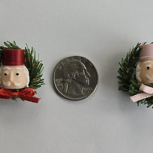 May include: Two miniature Christmas ornaments shaped like Santa Claus faces, each surrounded by a green wreath. One Santa wears a red top hat and bow tie, the other a pink top hat and bow. A quarter is between them.