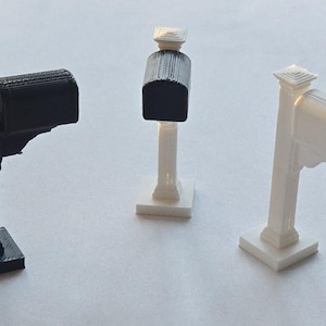 May include: Three miniature mailboxes in black and white. The mailboxes are mounted on posts with decorative details. The mailbox on the left is black, the middle one is black and white, and the one on the right is white.
