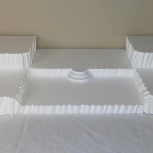May include: A white foam display stand featuring a multi-level tiered structure with flat platforms, vertical textured sides, two sets of small stairs on the outer edges, and a single small semi-circular step centered in the lower middle section.