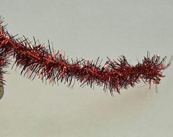 May include: A length of shiny red tinsel garland featuring thin, metallic, reflective strands that extend outward from a central wire core. The material has a bright, reflective finish that catches light, creating a textured, spiky appearance.