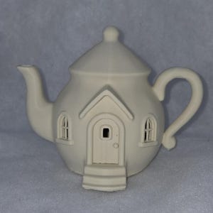Puede incluir: Una tetera de cerámica de color crema con forma de casa. La tetera presenta una puerta, ventanas y una parte superior en forma de chimenea. El asa y el pico son curvos. La tetera con forma de casa es un artículo decorativo único.