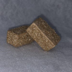 May include: Two small, square, brown blocks with a textured, fibrous appearance. The blocks are leaning against each other on a light gray surface. The blocks appear to be made of compressed plant material.