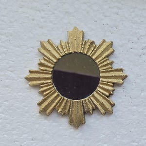 May include: A small circular mirror featuring a textured gold frame with radiating sunburst rays extending outward. The central reflective surface is dark and smooth, surrounded by a detailed, pointed metallic border that creates a star-like shape.