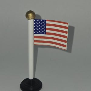 May include: A miniature American flag on a white pole with a gold-colored ball at the top. The flag features red and white stripes and a blue field with white stars. The base is black.
