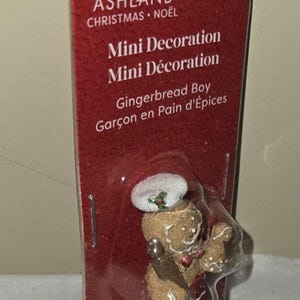 May include: A mini Christmas decoration featuring a gingerbread boy figurine. The figurine is brown with white icing details, wearing a white hat with a holly sprig. The packaging is red with white text.