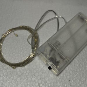 May include: A clear plastic battery pack with two slots and a small black switch is connected to a coil of silver wire fairy lights. The lights have small, warm white LED bulbs. The battery pack is rectangular.