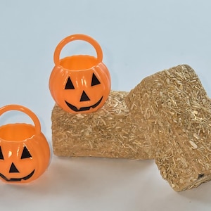 May include: Two orange plastic pumpkin-shaped treat buckets with black facial features and handles sit near a small hay bale. The Halloween-themed decorations are set against a white background.