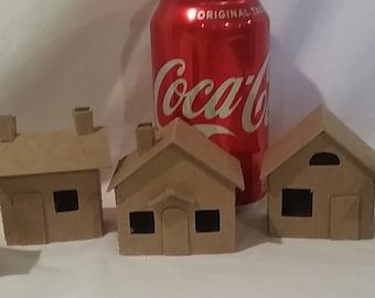 May include: Five miniature cardboard houses with brown paint, each with a chimney and windows. The houses are arranged in a row on a white surface, with a can of Coca-Cola in the background.