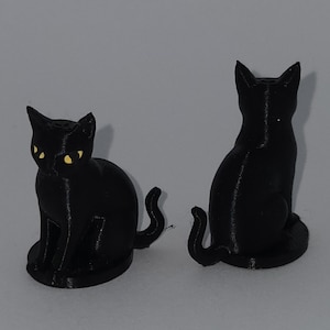 Op de afbeelding: Twee zwarte kattenbeeldjes. Eén kat kijkt naar voren met gele ogen, de andere is afgewend. Beide katten zitten met hun staart gekruld. De beeldjes staan op een effen achtergrond.