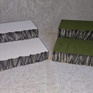 May include: Two-tiered display stands, one white and one green. The white stand has a textured surface and dark gray edges. The green stand has a grass-like surface and dark gray edges. Both stands are designed to elevate products.