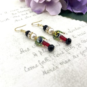 May include: Gold-coloured dangle earrings with black, red, and iridescent beads. Each earring features a black faceted bead, a white pearl, and a cluster of colourful beads. The earrings have a decorative design, suitable for special occasions.