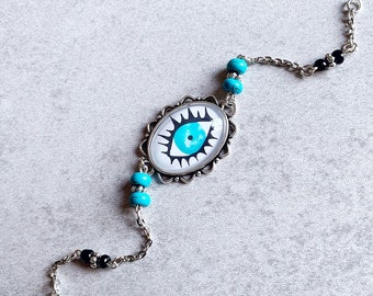 May include: A silver chain bracelet with a turquoise blue eye charm. The eye charm is surrounded by a silver frame with a black outline. The bracelet also has turquoise blue and black beads.