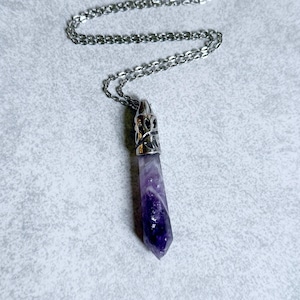 Pode incluir: Um colar com pingente de cristal de ametista roxa com uma corrente prateada. O cristal &eacute; de cor violeta profunda e &eacute; cortado em forma pontiaguda. O pingente tem uma tampa decorativa prateada.