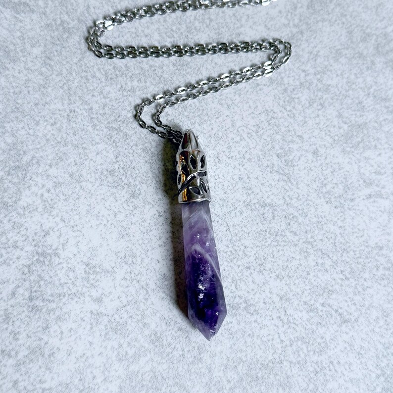 Pode incluir: Um colar com pingente de cristal de ametista roxa com uma corrente prateada. O cristal &eacute; de cor violeta profunda e &eacute; cortado em forma pontiaguda. O pingente tem uma tampa decorativa prateada.