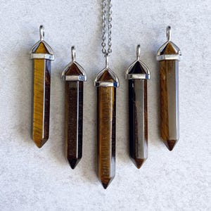 May include: Five gold tiger's eye crystal point pendants with silver caps. The pendants are arranged in a row, with the top pendant hanging from a silver chain.