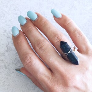 Könnte beinhalten: Eine Hand trägt einen Ring mit einem schwarzen Kristall, der in Silberdraht gewickelt ist. Die Hand ist mit hellblauem Nagellack lackiert.