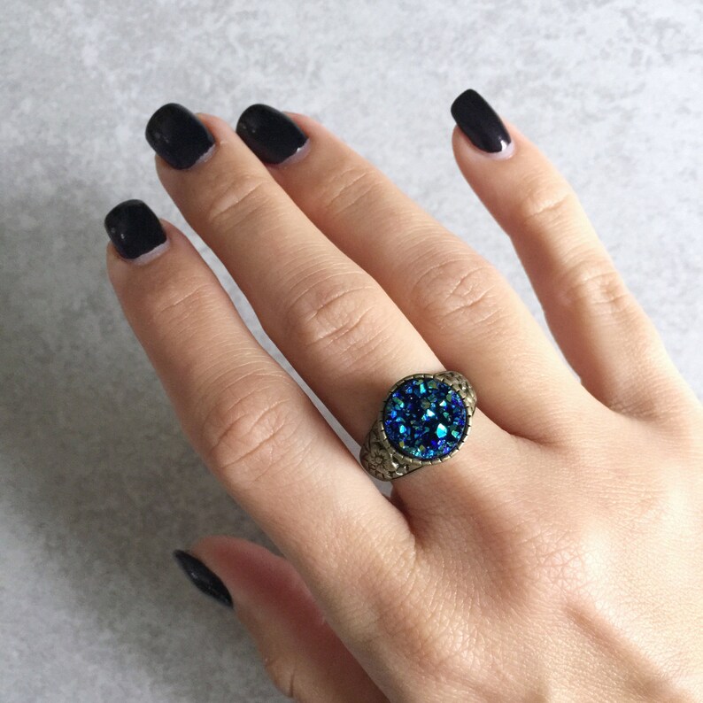 Peut inclure: Une bague avec une grande pierre pr&eacute;cieuse ronde bleue sertie dans une monture en m&eacute;tal dor&eacute;. La bague est port&eacute;e sur un doigt.