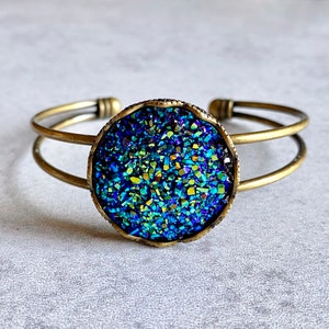 Puede incluir: Una pulsera de puño dorada con un gran centro redondo de piedra druzy azul y verde iridiscente. La piedra está engastada en un bisel dorado oscuro texturizado. La pulsera tiene un diseño de doble banda.