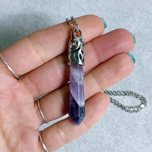Pode incluir: Colar com pingente de cristal roxo e branco com corrente prateada. O cristal tem formato hexagonal com uma tampa de metal prateado. O colar &eacute; exibido em um fundo cinza claro.