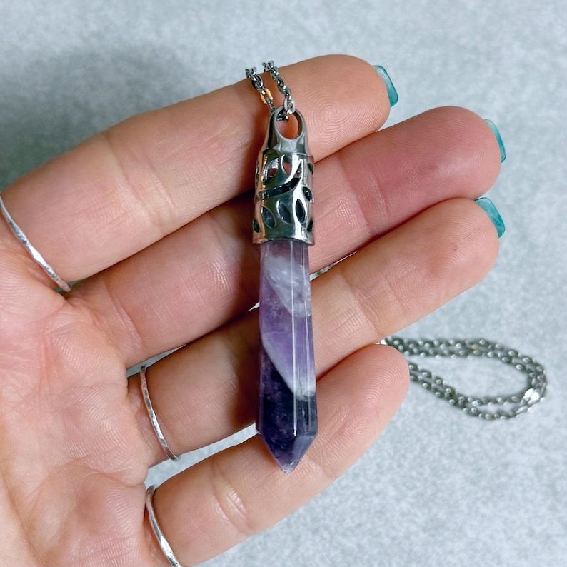 Pode incluir: Colar com pingente de cristal roxo e branco com corrente prateada. O cristal tem formato hexagonal com uma tampa de metal prateado. O colar &eacute; exibido em um fundo cinza claro.