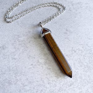 May include: A silver chain necklace with a brown tiger's eye crystal pendant. The pendant is a long, pointed shape with a silver cap at the top.
