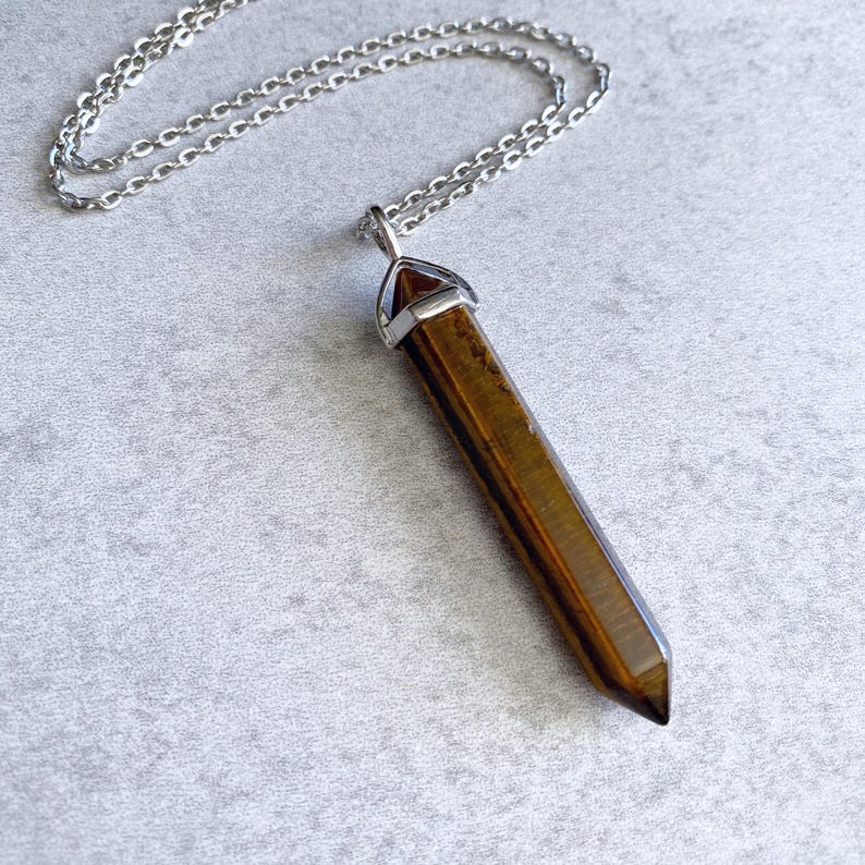 May include: A silver chain necklace with a brown tiger's eye crystal pendant. The pendant is a long, pointed shape with a silver cap at the top.