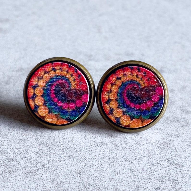 May include: A pair of round stud earrings with a colorful tie-dye design. The earrings feature a spiral pattern in shades of red, orange, yellow, blue, and purple. The earrings have a bronze-colored metal border and are set against a neutral background.