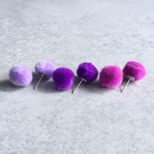 Puede incluir: Cinco pares de pendientes de pomp&oacute;n de color lila y lavanda. Los pendientes est&aacute;n dispuestos en fila del m&aacute;s claro al m&aacute;s oscuro.