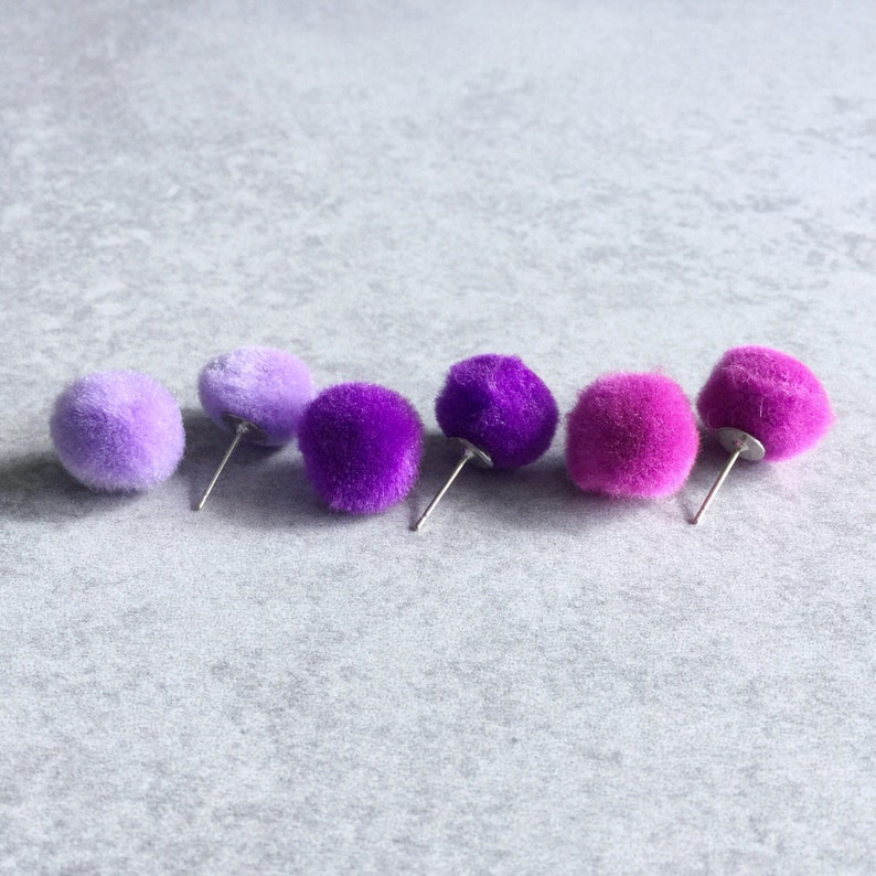 Puede incluir: Cinco pares de pendientes de pomp&oacute;n de color lila y lavanda. Los pendientes est&aacute;n dispuestos en fila del m&aacute;s claro al m&aacute;s oscuro.