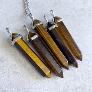 May include: Five gold tiger's eye crystal point pendants with silver caps and loops. The pendants are arranged in a row, with the largest pendant at the top and the smallest pendant at the bottom.