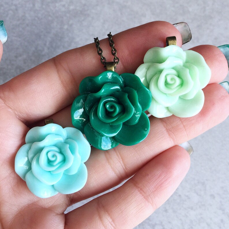 Puede incluir: Tres dijes de resina con forma de rosa en tonos verdes y azul claro. Los dijes est&aacute;n unidos a un collar de cadena plateada.