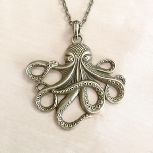 Könnte beinhalten: Halskette mit einem bronzefarbenen Oktopus-Anhänger. Der Oktopus hat detaillierte Tentakel mit Saugnäpfen und einen strukturierten Kopf. Die Halskette ist ebenfalls bronzefarben. Ein Schmuckstück mit maritimem Thema.