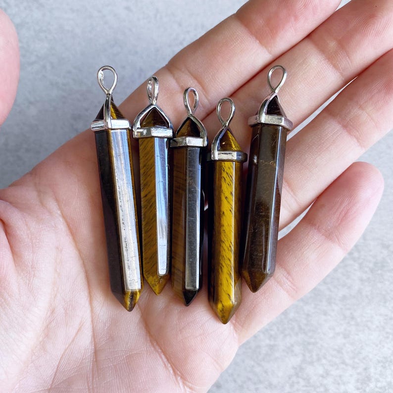 May include: Five gold tiger's eye gemstone pendants with silver caps and loops. The pendants are in the shape of a point and are arranged in a row on a hand.