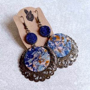 May include: A pair of dangle earrings with blue and gold marbled resin and a filigree design. The earrings are on a brown card with a crystal and the letter B.